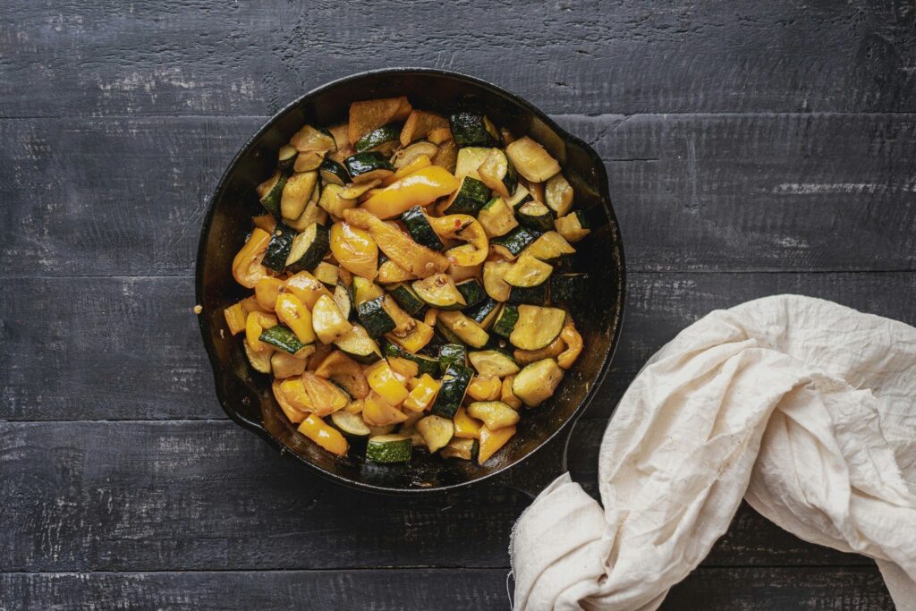 Poêlée de courgettes et de poivrons jaunes dans une poêle en fonte, vue de dessus sur une table en bois sombre
